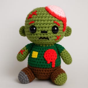 Könnte beinhalten: Handgefertigte Häkelpuppe eines Zombies mit grünem Körper, dunkelgrünem Shirt und brauner Hose. Die Puppe hat eine rote Wunde am Kopf und an der Brust, mit roten Stichen und einem gelben Fleck. Schwarze Augen und ein gestickter Mund.