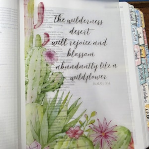 May include: A translucent vellum insert placed over a Bible page, featuring a watercolor illustration of green cacti and pink flowers on the left and bottom. The text reads: the wilderness desert will rejoice and blossom abundantly like a wildflower ISAIAH 35:1.