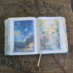 May include: An open Bible with colorful index tabs along the edges. The left page features a blue-toned illustration of a night scene, while the right page displays a watercolor-style image of a baptism in water with a white dove descending from above.