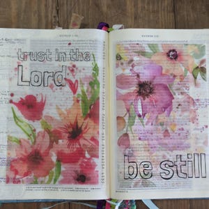 以下が含まれることがあります： 水彩画のフローラルアートと、黒いアウトライン文字で「trust in the Lord」と「be still」というフレーズが書かれた開いた聖書のページ。花はピンク、赤、オレンジの色合いです。