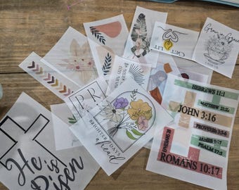 May include: A collection of translucent vellum sheets featuring various printed designs including floral bouquets, crosses, and religious text such as He is Risen, Grow in Grace, and various Bible verses like John 3:16 and Romans 10:17 in black and color.