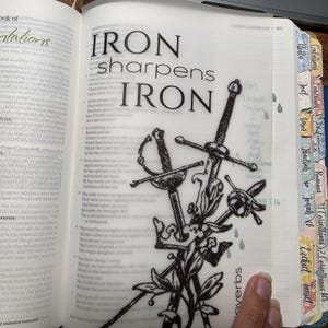 May include: A translucent vellum page insert in a book featuring the black printed text IRON sharpens IRON above a detailed black ink illustration of three crossed swords and a crown, with colorful index tabs visible on the right edge of the book.