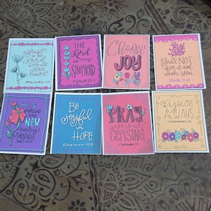 Op de afbeelding: Acht rechthoekige kaarten met kleurrijke illustraties en handgeschreven bijbelverzen. De ontwerpen bevatten bloemmotieven en inspirerende teksten zoals Trust in the Lord, The Lord is my Shepherd, Choose Joy, Fear Not, His mercies are new, Be joyful in hope, Pray without ceasing en Rejoice Always.