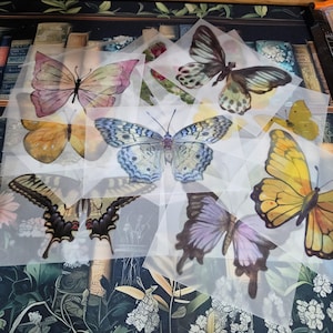May include: A collection of watercolor butterfly designs in various colors, including yellow, purple, and blue, printed on translucent paper. The butterflies have detailed wing patterns and are arranged on a floral and book-themed background.