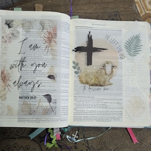 May include: An open Bible featuring translucent vellum inserts. The left page displays the text "I am with you always" and "MATTHEW 28:20" with botanical illustrations. The right page shows a sheep, a cross, and the text "HE LEFT THE 99 to rescue me."