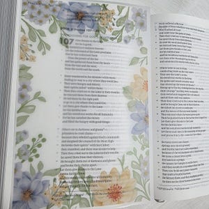 May include: Open Bible with text from Psalms 106-107. The pages are open to a passage with the heading "Giving for God's Deliverance." Floral watercolor design in pastel colors adorns the left page. The book is on a light-colored surface.