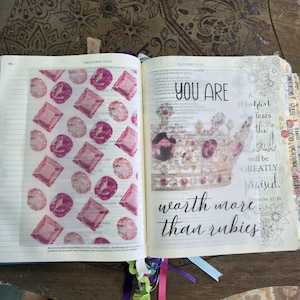 May include: An open book with a translucent overlay featuring a pattern of pink rubies on the left page. The right page displays a gold crown illustration with the text YOU ARE worth more than rubies and a floral design on the right margin.