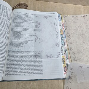 May include: An open Bible with thin, translucent vellum pages featuring a delicate line-art floral pattern. The book includes colorful, labeled index tabs along the side and a brown ribbon bookmark. Additional loose vellum sheets with the same floral design are visible.