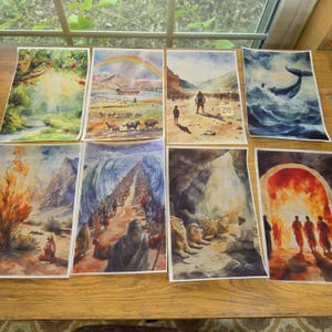 May include: A collection of eight rectangular watercolor prints depicting various biblical scenes, including a lush garden, a rainbow over an ark, a desert landscape, a whale in the ocean, a burning bush, a parted sea, lions in a den, and a fiery furnace.