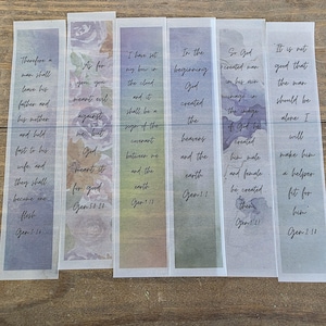 May include: Set of six rectangular bookmarks with Bible verses printed in black script. Each bookmark features a watercolor background with floral accents in shades of purple, blue, and green. The bookmarks are arranged on a wooden surface.