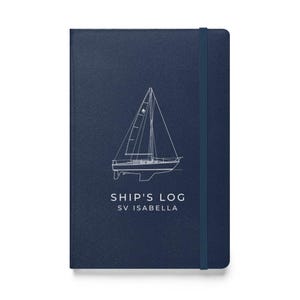 May include: A dark blue hardcover notebook with a vertical elastic closure band. The front cover features a white line drawing of a sailboat centered above the text "SHIP'S LOG" and "SV ISABELLA" printed in a clean, sans-serif white font.