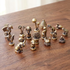 Könnte beinhalten: Eine Sammlung kleiner Messingfiguren im Antik-Stil, die verschiedene Tiere, Pflanzen und Objekte darstellen. Das Set enthält einen Drachen, eine Fledermaus, eine Schnecke, einen Hasen, eine Katze, einen Dinosaurier, einen Frosch, einen Vogel und einen Bienenstock.