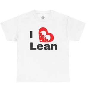 May include: White t-shirt featuring the text "I Lean" in black, with a red heart-shaped graphic. The heart graphic has two white eyes with red pupils. The shirt has a crew neck and short sleeves.