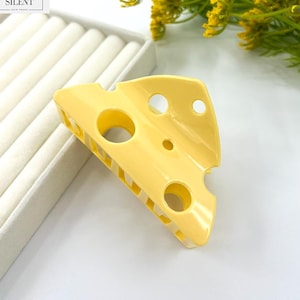May include: A yellow cheese-shaped hair clip with several round holes. The clip has a claw-like design and the word "SILENT" is printed on a white label in the upper left corner. The clip is resting on a white surface.