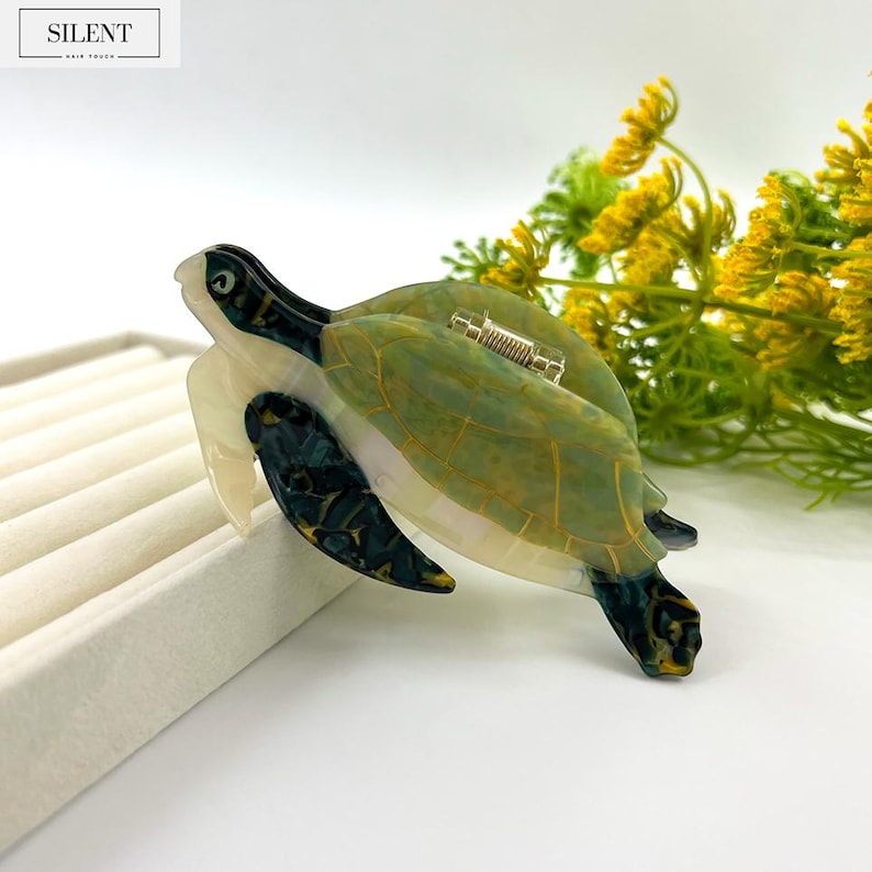May include: A sea turtle-shaped hair clip in shades of green, cream, and black. The clip features a detailed design with a metallic clasp. The word "SILENT" is printed in a white box at the top left.