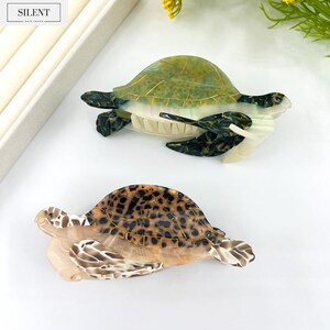 May include: Two decorative hair clips shaped like sea turtles. One clip is green and white, the other is brown and tan with a tortoise shell pattern. The clips are made of a translucent material and have a glossy finish. The word "SILENT" is in the top left corner.