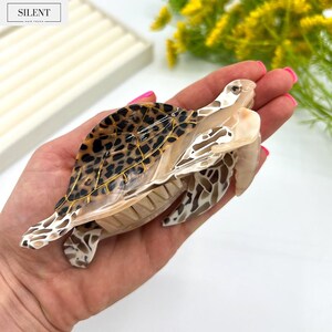 May include: A decorative turtle hair claw clip with a tortoiseshell pattern on the shell and gold accents. The clip is beige and brown, with the word "SILENT" in a white box in the upper left corner.
