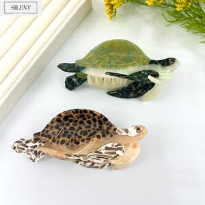 May include: Two decorative turtle hair clips. One clip features a green and gold shell, while the other has a brown and black spotted shell. The clips are made of a translucent material and are set against a white background. The brand name "SILENT" is visible.
