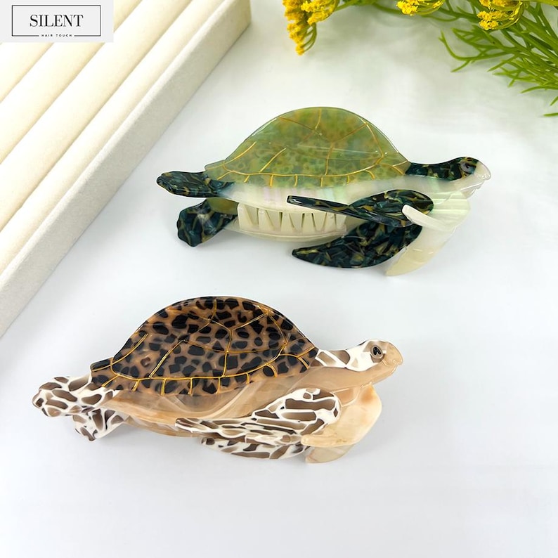 May include: Two decorative turtle hair clips. One clip features a green and gold shell, while the other has a brown and black spotted shell. The clips are made of a translucent material and are set against a white background. The brand name "SILENT" is visible.