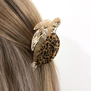 May include: A tortoise-shaped hair clip made of tortoiseshell and leopard print acrylic. The hair accessory is attached to blonde hair. The word "SILENT" is printed in a rectangular box in the upper right corner.