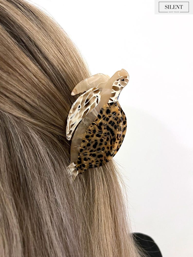 May include: A tortoise-shaped hair clip made of tortoiseshell and leopard print acrylic. The hair accessory is attached to blonde hair. The word "SILENT" is printed in a rectangular box in the upper right corner.