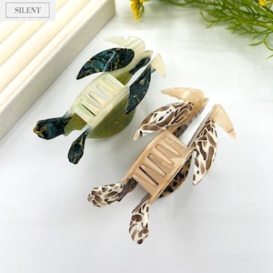 May include: Two decorative hair clips shaped like turtles. One clip is green and black, the other is tan and brown. The clips have a comb-like design and are made of a glossy material. The word "SILENT" is printed on a white label.