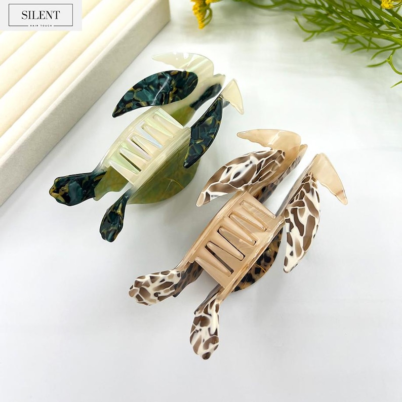 May include: Two decorative hair clips shaped like turtles. One clip is green and black, the other is tan and brown. The clips have a comb-like design and are made of a glossy material. The word "SILENT" is printed on a white label.