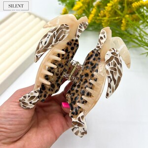 May include: A large hair claw clip with a tortoiseshell and leopard print design. The clip features a combination of tan, brown, and cream colors. The clip is made of plastic and has a strong grip for holding hair in place. The brand name "SILENT" is visible in the top left corner.