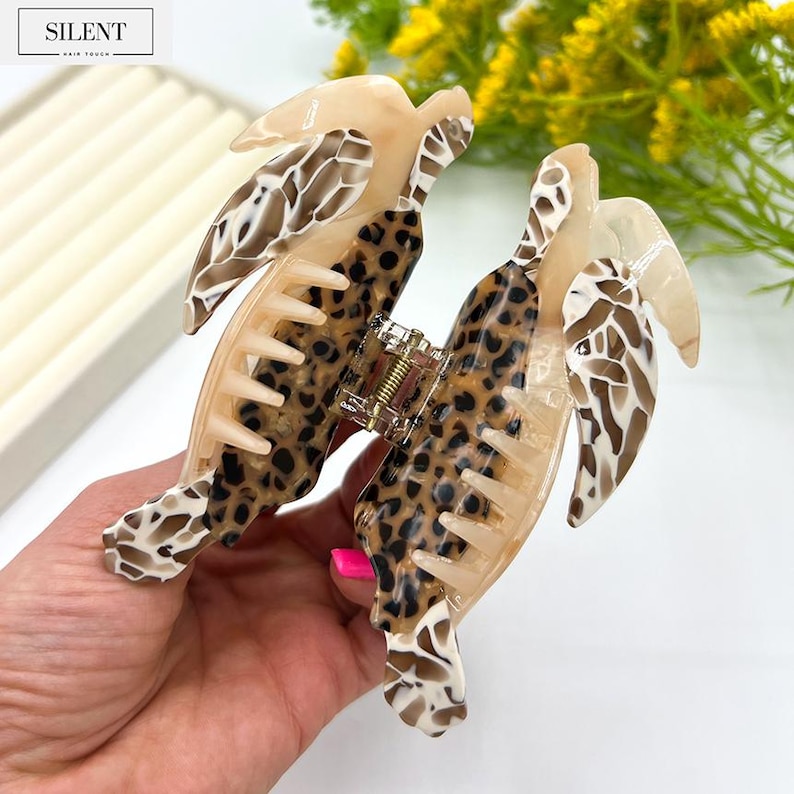 May include: A large hair claw clip with a tortoiseshell and leopard print design. The clip features a combination of tan, brown, and cream colors. The clip is made of plastic and has a strong grip for holding hair in place. The brand name "SILENT" is visible in the top left corner.