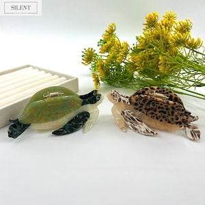 May include: Two turtle-shaped hair clips. One is green and white, the other is tan with brown spots. Both have a gold-colored metal clasp. The background includes yellow flowers and a white surface. The top left corner has the text "SILENT HAIR TOUCH".
