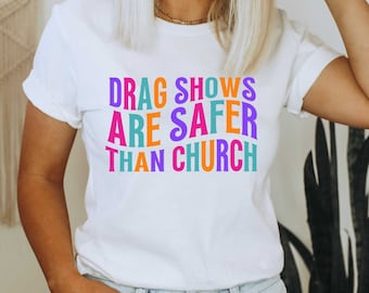 May include: A white t-shirt with the text "DRAG SHOWS ARE SAFER THAN CHURCH" printed in rainbow colours.