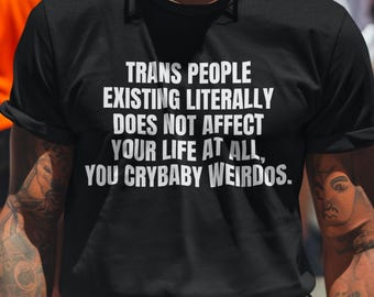 May include: Black t-shirt with the text "TRANS PEOPLE EXISTING LITERALLY DOES NOT AFFECT YOUR LIFE AT ALL, YOU CRYBABY WEIRDOS." The image shows a person wearing the shirt in a crowd with rainbow flags.