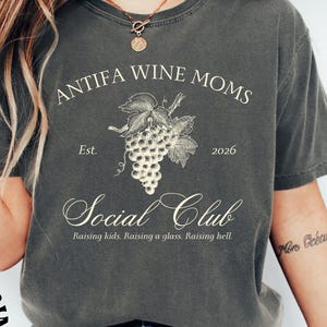 May include: Dark gray t-shirt with the text "ANTIFA WINE MOMS" above a grape illustration and "Social Club" below. The shirt also includes the text "Raising kids. Raising a glass. Raising hell."