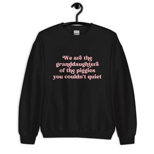 Pode incluir: Camisola preta com o texto "We are the granddaughters of the piggies you couldn't quiet" em letras cor-de-rosa. A camisola est&aacute; pendurada num cabide preto.