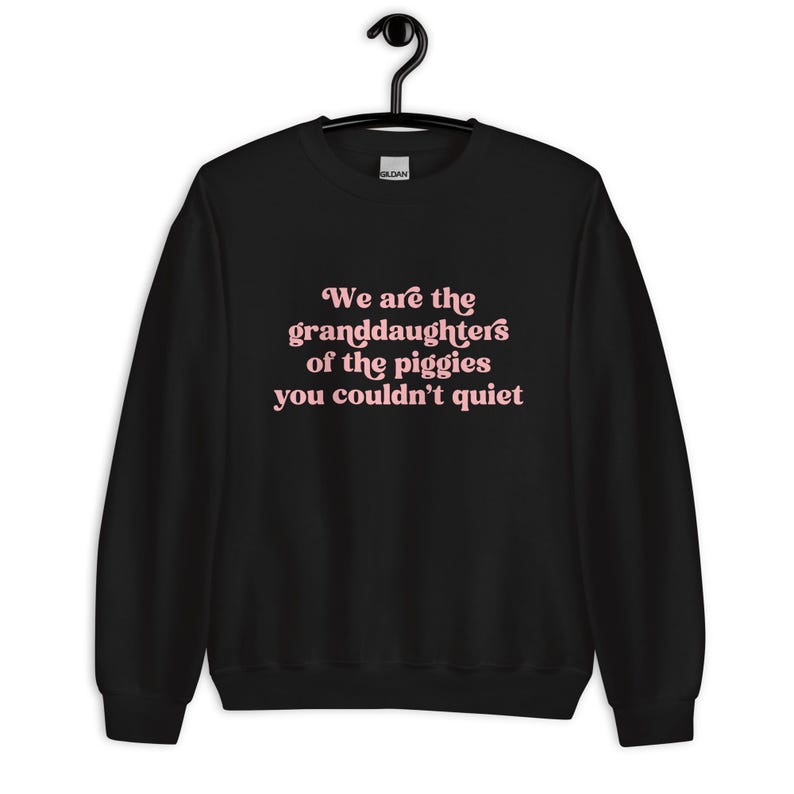 Pode incluir: Camisola preta com o texto "We are the granddaughters of the piggies you couldn't quiet" em letras cor-de-rosa. A camisola est&aacute; pendurada num cabide preto.