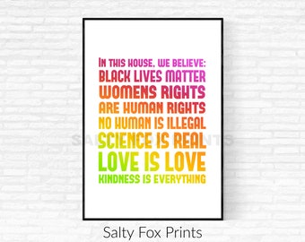 May include: A white printable poster with a rainbow gradient of text that reads "In this house, we believe: Black Lives Matter, Women's Rights are Human Rights, No Human is Illegal, Science is Real, Love is Love, Kindness is Everything". The poster is framed in a black frame and is hanging on a white brick wall. The text "Salty Fox Prints" is printed below the poster.