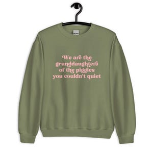 Pode incluir: Camisola verde-oliva com o texto rosa: "We are the granddaughters of the piggies you couldn't quiet". A pe&ccedil;a de roupa est&aacute; pendurada num cabide preto.