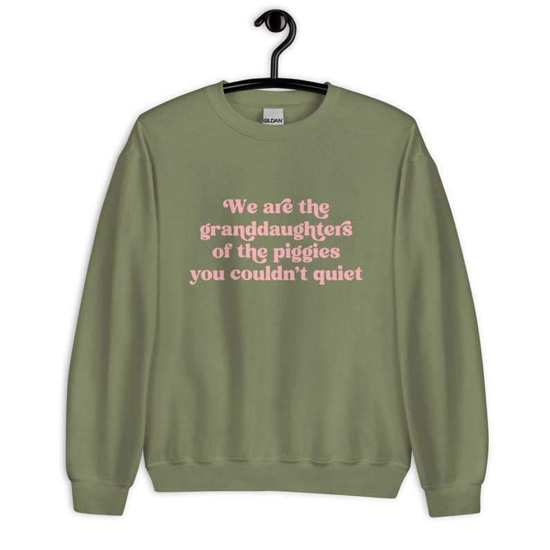 Pode incluir: Camisola verde-oliva com o texto rosa: "We are the granddaughters of the piggies you couldn't quiet". A pe&ccedil;a de roupa est&aacute; pendurada num cabide preto.