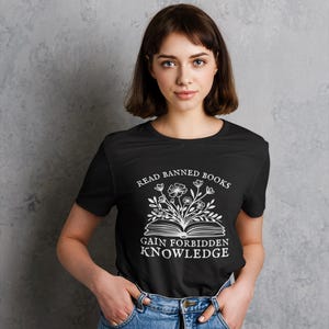 May include: Black t-shirt with the text "READ BANNED BOOKS GAIN FORBIDDEN KNOWLEDGE" in white. The design includes an open book with flowers and leaves. The person is wearing blue jeans.