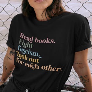 K&ouml;nnte beinhalten: Schwarzes T-Shirt mit dem Text "Read books. Fight fascism. Look out for each other." in bunten Buchstaben.