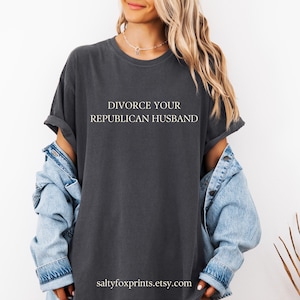 Puede incluir: Una camiseta gris oscuro con el texto "DIVORCE YOUR REPUBLICAN HUSBAND" impreso en blanco en la parte delantera. La camiseta la lleva puesta una persona con el pelo largo rubio y una chaqueta vaquera.