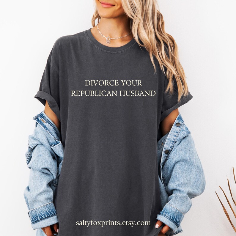 Puede incluir: Una camiseta gris oscuro con el texto "DIVORCE YOUR REPUBLICAN HUSBAND" impreso en blanco en la parte delantera. La camiseta la lleva puesta una persona con el pelo largo rubio y una chaqueta vaquera.
