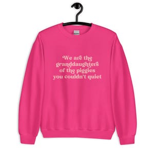 Pode incluir: Camisola rosa choque com a frase "We are the granddaughters of the piggies you couldn't quiet" em rosa claro. A camisola est&aacute; pendurada num cabide preto.