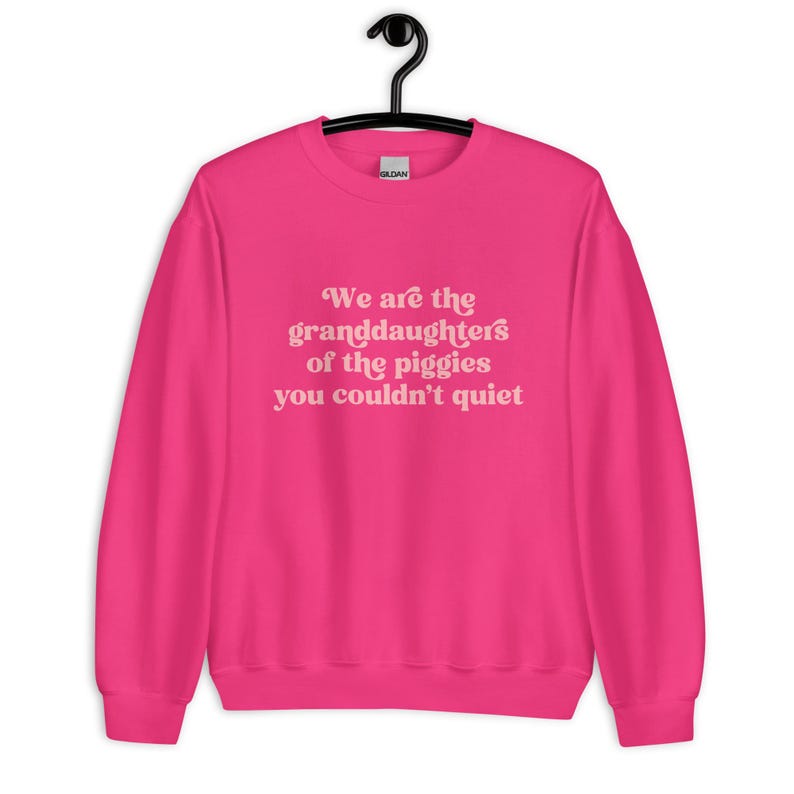 Pode incluir: Camisola rosa choque com a frase "We are the granddaughters of the piggies you couldn't quiet" em rosa claro. A camisola est&aacute; pendurada num cabide preto.