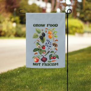 May include: A blue garden flag with a white background and a colorful illustration of various fruits and vegetables. The text "Grow Food Not Fascism" is printed in green letters.