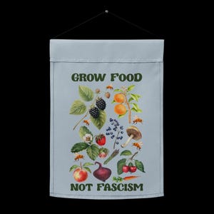 May include: A blue banner with the text "GROW FOOD NOT FASCISM" in green letters. The banner is decorated with a variety of fruits, vegetables, and mushrooms, including blackberries, oranges, strawberries, cherries, and a mushroom.