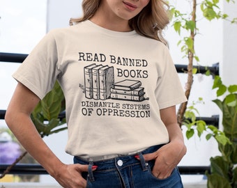 Puede incluir: Una camiseta blanca con un gráfico negro que dice "Leer libros prohibidos Desmantelar sistemas de opresión". El gráfico presenta una pila de libros.