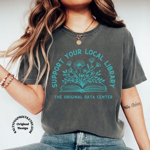 Puede incluir: Una camiseta gris carbón con un gráfico en color turquesa de un libro abierto del que brotan flores. El texto "SUPPORT YOUR LOCAL LIBRARY" aparece en arco sobre el libro y "THE ORIGINAL DATA CENTER" está impreso debajo del libro.