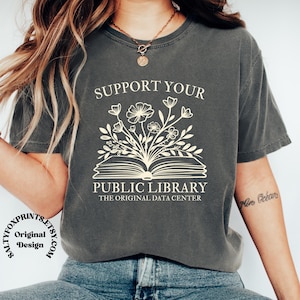 May include: A charcoal gray short-sleeve t-shirt featuring a cream-colored graphic of an open book with flowers growing from the pages. The text reads SUPPORT YOUR PUBLIC LIBRARY THE ORIGINAL DATA CENTER in a clean, sans-serif font style.
