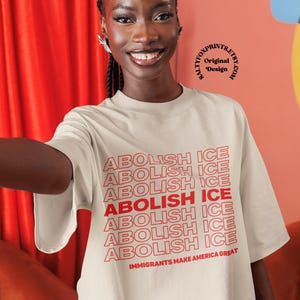 K&ouml;nnte beinhalten: Ein cremefarbenes T-Shirt mit dem Text "ABOLISH ICE" in Rot wiederholt und vertikal gestapelt. Der Satz "IMMIGRANTS MAKE AMERICA GREAT" ist darunter gedruckt. Das Bild zeigt eine l&auml;chelnde Person.